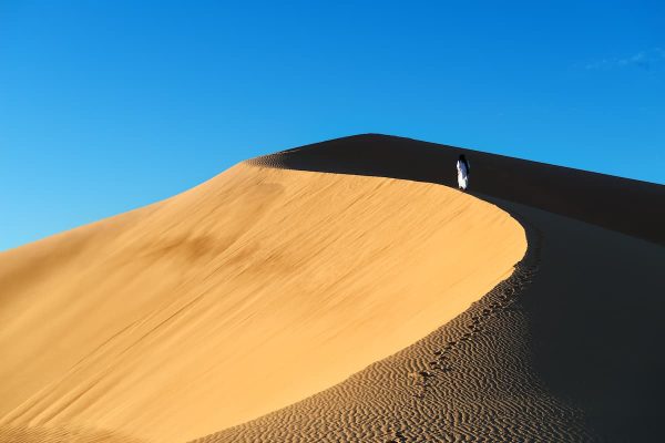 A traditional dressed moroccan man walks up a sand dune, Sahara desert, Morocco.